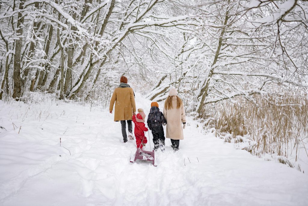 Zwei erwachsene und zwei Kinder laufen mit einem Schlitten in einem verschneiten Wald auf einem Wanderweg