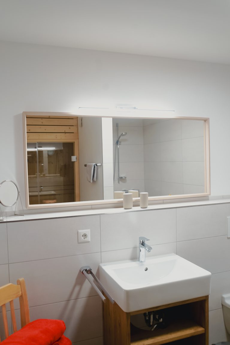 Ein großer Spiegel über einem Waschbecken, darin reflektiert sich eine Sauna und die Dusche