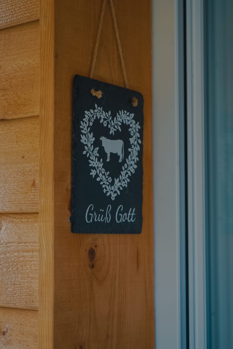 Ein Schild an einer Holzwand, auf dem grüß Gott steht und eine Kuh zu sehen ist.