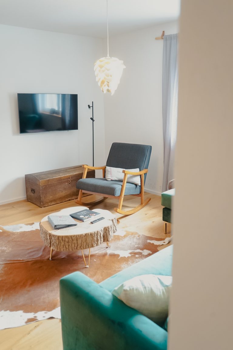Ein an der Wand hängender Fernseher, davor steht ein Schaukelstuhl und ein Holztisch, auf dem Boden ein Kuhfell-Teppich.