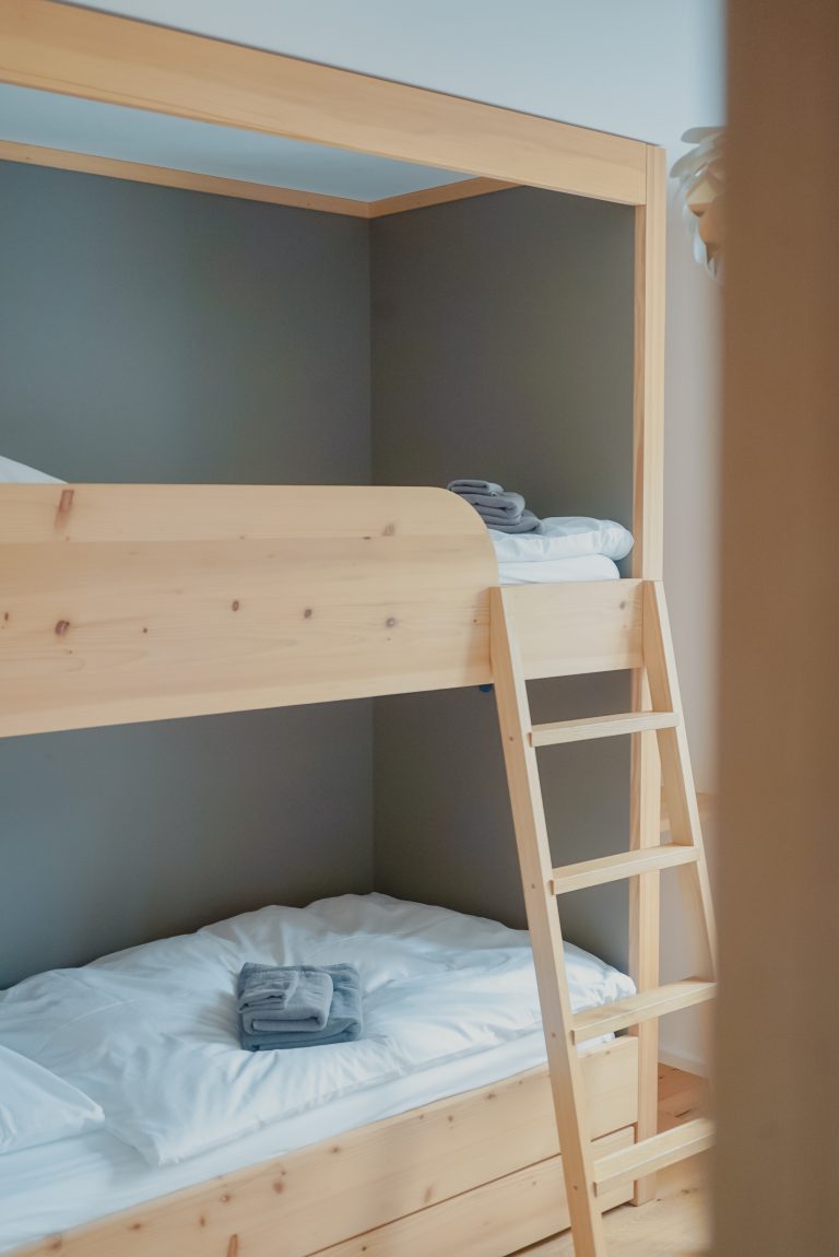 Zwei hölzerne Stockbetten mit einer Leiter um in das obere Bett zu steigen, im Hintergrund eine graue Wand.